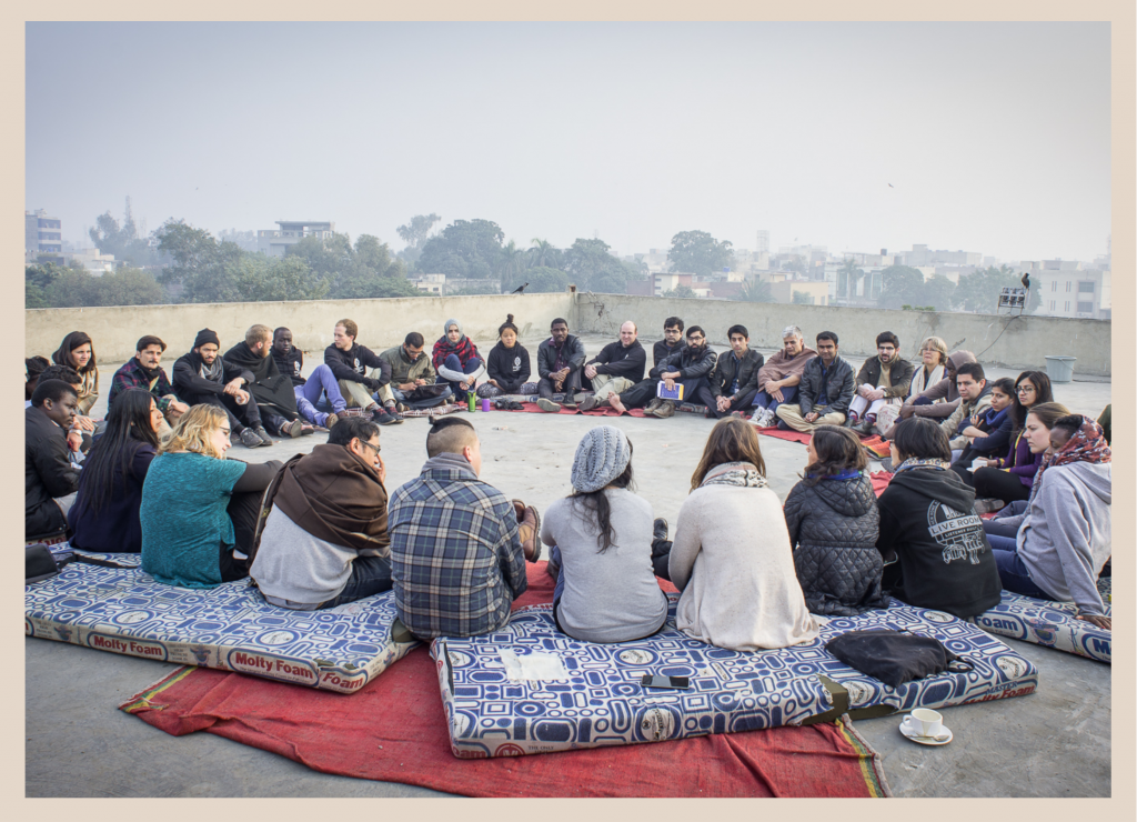 Image showing a large group of people from diverse backgrounds sitting in a circle on blue and white mats laid out on the rooftop of a building in Lahore, Pakistan.  