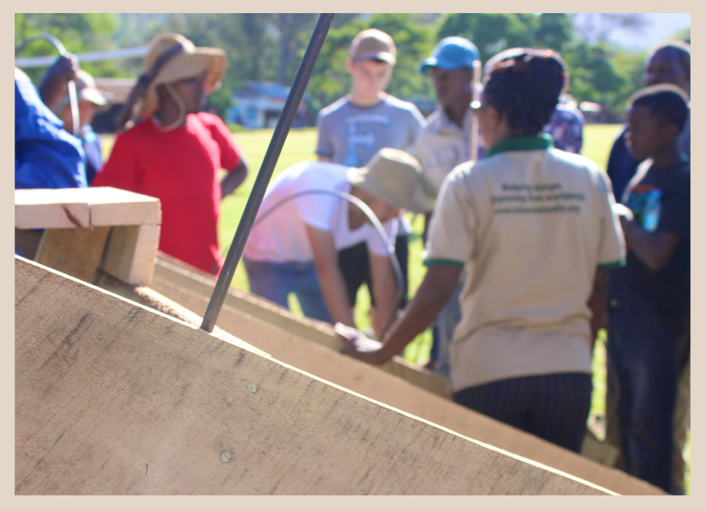 An image with a wooden prototype in the foreground and a group of people engaged in conversation in the background. The group includes a team of MIT students, workers from a local NGO, and members of the town of Leguruki, Tanzania.  